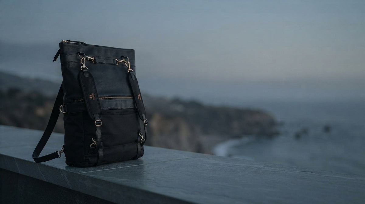 A black convertible backpack purse sits on a stone ledge in a coastal setting with a clear, open sky in the center.
